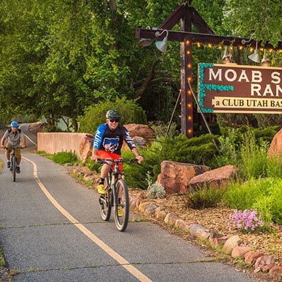 Two male bicyclists on a bike path riding past the rectangular Moab Springs Ranch sign.