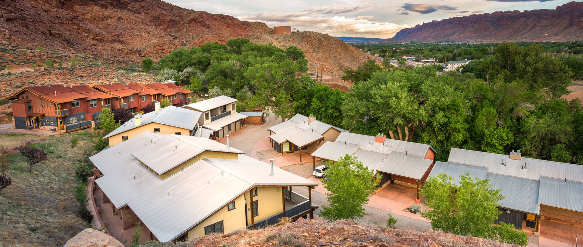 Aerial view of rows of the triangular roofs of townhouse units on Moab Springs Ranch with tall green trees and grasslands, and a rural road leading into the property.