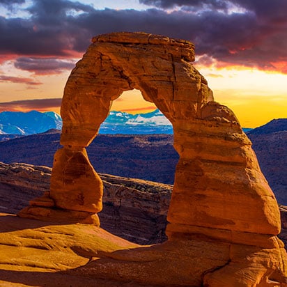 An arch structure of sandstone stands against the background of La Sal Mountains in the glow of sunset at Arches National Park in Utah.