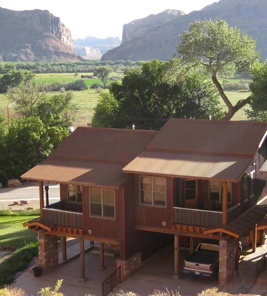 A view of two attached bungalow Units on Moab Springs Ranch, each with a two-car garage, standing amongst tall green trees and towering cliffs in the background.
