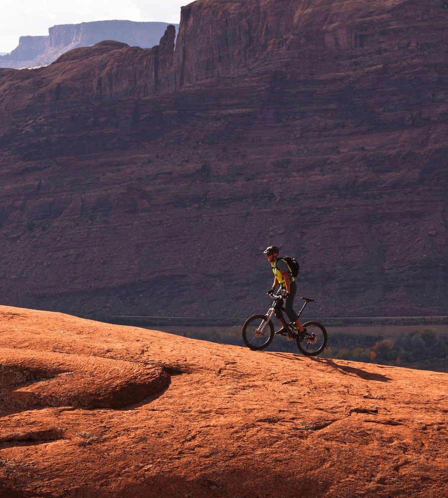 A male mountain biker is riding up a biking trail against the backdrop of a towering sandstone cliff in Moab, Utah.