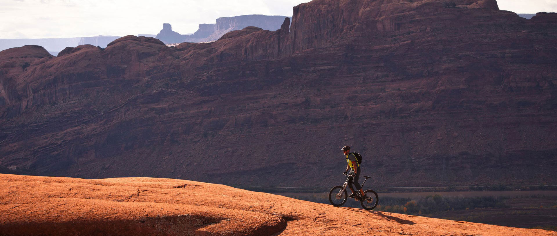 A large panoramic view of the monolithic mountains and sandcliffs of Moab, Utah and a lone male mountain biker riding up a trail.