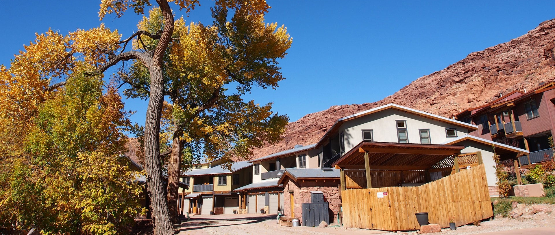 Tall majestic trees bearing orange and yellow leaves tower above triangular shaped roofs of townhomes at Moab Springs Ranch under bright blue skies.